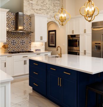 Elegant white kitchen with navy island, gold pendant lights, marble countertops, and patterned backsplash